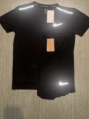 Nike Miller set - Säljer ett svart T-shirt och shorts från Nike med t-shirt och shorts. Båda plaggen har reflekterande detaljer och Nike-logga. Perfekt för löpning eller gym. Materialet är lätt och andas, designen är enkel och sportig.STORLEKSINFO: Lappen säger L (Kina), men motsvarar en EU Small (S). • Bröst: 95–101 cm. • Midja: 88–94 cm.