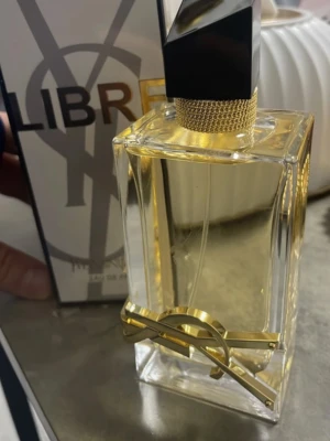 Yves Saint Laurent Libre Eau de Parfum - Stilren parfymflaska från Yves Saint Laurent, Libre Eau de Parfum. Flaskan är rektangulär i genomskinligt glas med gulddetaljer och svart asymmetrisk kork. Den har en lyxig känsla med YSL-loggan i guld på framsidan.
