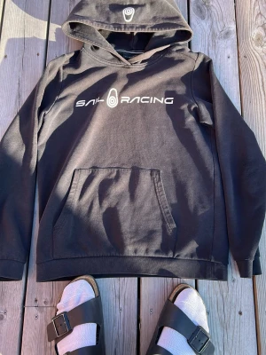 Svart Sail Racing hoodie med tryck - Svart hoodie från Sail Racing med vitt logotryck på bröstet och en mindre logga på huvan. Klassisk känguruficka framtill och mjukt material som känns skönt mot huden. Perfekt för dig som gillar en clean och sportig stil.