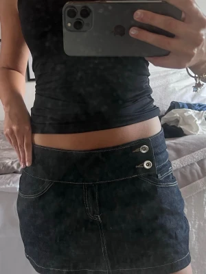 Lågmidjad blå jeanskjol med knappar - Snygg lågmidjad marinblå jeanskjol med vita kontrastsömmar och två silverfärgade knappar framtill. fickor bak med knappdetalj och sitter lågt på höften. Inbyggda shorts. Från bershka i stl 36 
