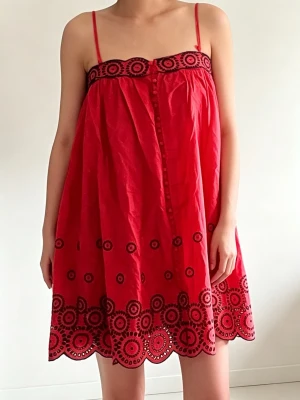 Röd kort klänning med broderi - Söt röd kort klänning med tunna axelband och dekorativa svarta broderier längs överdelen och nederkanten. Klänningen har en luftig passform och knappar framtill. Perfekt för varma dagar och ger en lekfull vibe.