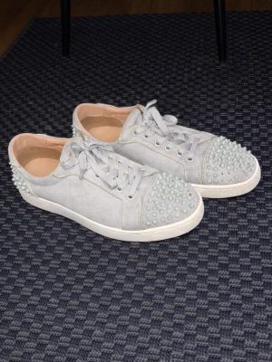 Gråa sneakers med nitar från Louboutin - Unika gråa sneakers från Christian Louboutin med coola nitar på tå och häl. Skorna har en låg silhuett, snörning och är gjorda i mocka med vit sula. Perfekta för dig som vill sticka ut med din stil.