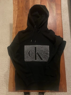 Svart hoodie från Calvin Klein Jeans - Säljer en svart hoodie från Calvin Klein Jeans med stor logotyp på bröstet i grått och svart. Hoodien har dragsko i huvan och ribbade muddar vid ärmslut och nederkant. Perfekt för dig som gillar stilrena och klassiska plagg.