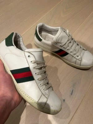 Gucci vita sneakers med röd/grön rand - Klassiska vita sneakers från Gucci i läder med den ikoniska gröna och röda randen på sidan. Skorna har lågt skaft, vita snören och grön detalj på hälen. Perfekta för dig som vill ha en stilren och lyxig look.