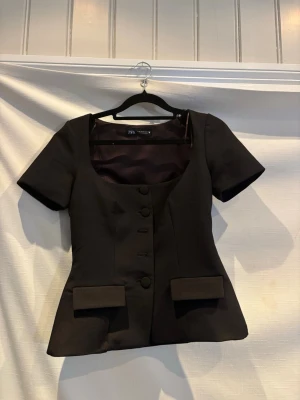 Svart kavaj med kort ärm från Zara - Stilren svart kavaj från Zara med korta ärmar och markerad midja. Kavajen har fyra klädda knappar framtill, två dekorativa ficklock och en djup, rundad urringning. Materialet är slätt och ger en elegant look. Perfekt för dig som vill ha en modern twist på klassisk kavaj.