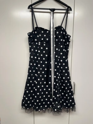 Svart prickig kort klänning - Snygg svart kort klänning med vita prickar och tunna axelband. Klänningen har en söt volangkant i tyll längst ner och ett band som kan knytas i nacken. Perfekt för dig som gillar retrostil och vill sticka ut.