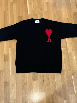 Svart stickad tröja från Ami Paris storlek M/L - Svart stickad tröja från Ami Paris med röd hjärtlogga och bokstaven 'A' på bröstet. Tröjan har rund halsringning och ribbade muddar vid ärmslut och nederkant. Perfekt för dig som gillar stilrena plagg med en twist. Passar M-L.