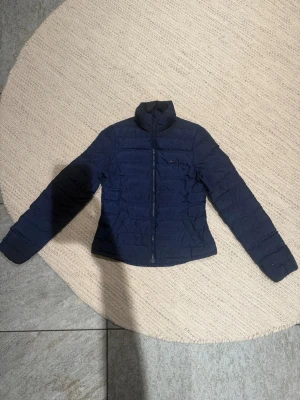 Mörkblå pufferjacka från Tommy Hilfiger - Snygg mörkblå pufferjacka från Tommy Hilfiger med diskret logga på bröstet. Jackan har hög krage, dragkedja framtill och quiltad design. Perfekt för kyliga dagar och enkel att matcha med olika outfits. Finns två st, storlek 36 och 38