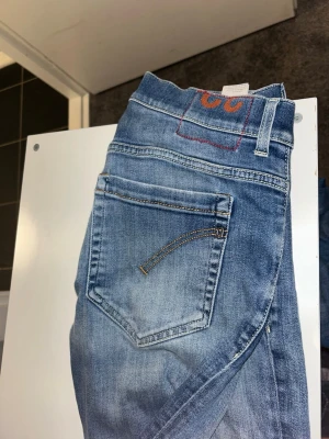 Blå jeans med slitningar - Säljer ett par klassiska blå jeans med snygga slitningar och tvättad look. Jeansen har fem fickor, bälteshällor och knappgylf. Passar perfekt till en avslappnad stil och har en rak passform. Märke syns ej tydligt men de har en röd detalj på bakfickan. W32 sitter som 30,29. Kortare i längden 