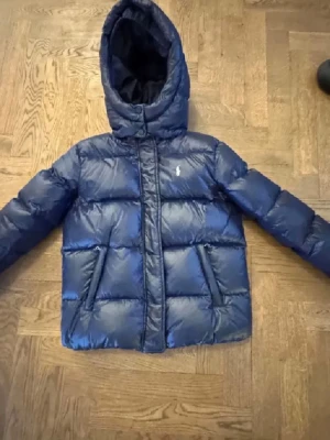 Blå dunjacka från Polo Ralph Lauren - Snygg blå dunjacka från Polo Ralph Lauren med huva och dragkedja framtill. Jackan har två fickor med dragkedja och den klassiska loggan broderad på bröstet. Perfekt för kalla vinterdagar tack vare det tjocka dunfodret och den puffiga designen. Barn storlek i L 