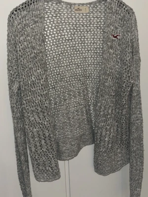 Grå stickad cardigan från Hollister - Snygg gråmelerad stickad cardigan från Hollister med öppen front och lång ärm. Tröjan har ett hålmönster och en liten vinröd Hollister-logga broderad på bröstet. Perfekt att slänga över en topp för en avslappnad look.