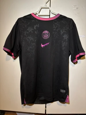 Svart PSG matchtröja med rosa detaljer - Snygg svart PSG matchtröja från Nike med diskret blommönster i tyget, rosa detaljer vid krage och ärmslut samt rosa PSG-logga och Nike-märke på bröstet. Stort vitt nummer 10 på ryggen. Perfekt för fotboll eller streetwear.