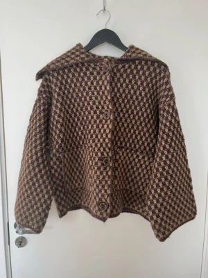 Brunrutigt retroinspirerad kappa - Säljer en brun och beige kappa med rutigt mönster och stora knappar framtill. Kappan har en bred krage och är tillverkad i ett tjockare, stickat material som ger en vintagekänsla. Perfekt för dig som gillar unika plagg med retrovibbar.