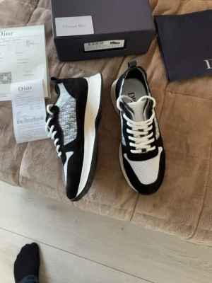 Dior skor - Snygga svartvita sneakers från Dior med klassisk logga på sidan, meshdetaljer och mockaliknande partier. Tjock vit sula med svarta gummiprickar undertill och vita skosnören. Perfekta för dig som vill ha en exklusiv och trendig look.