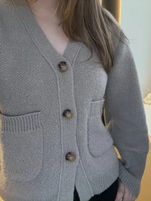 Beige stickad kofta med stora knappar - Mysig beige stickad kofta med v-ringning och tre stora bruna knappar framtill. Koftan har två framfickor och ribbade muddar vid ärmslut och nederkant. Perfekt för lager på lager och enkel att matcha med olika outfits. Knappt använd, lite nopprig men inget som märks av