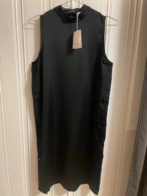 Svart ärmlös klänning från Samsoe & Samsoe - Stilren svart ärmlös klänning med hög hals och knappar längs sidorna. Klänningen har rak passform och är tillverkad i ett mjukt, slätt material som ger en elegant look. Perfekt för dig som gillar minimalistisk stil.
