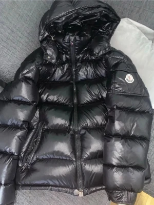 Svart dunjacka från Moncler - Snygg svart dunjacka från Moncler med glansig finish och klassisk logga på ärmen. Jackan har huva, dragkedja framtill och två sidofickor med dragkedja. Perfekt för kalla vinterdagar och riktigt varm tack vare dunfyllningen. Går att scanna på ärmen och påse, tags allt ingår