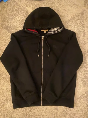 Svart hoodie från Burberry - Svart hoodie från Burberry med dragkedja framtill, klassisk huva med snören och ikoniskt rutigt foder i huvan. Broderad Burberry-logga på bröstet. Perfekt för en stilren och avslappnad look.