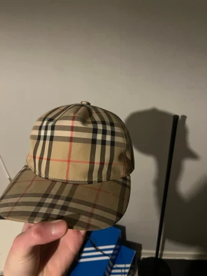 Burberry beige rutig keps - Säljer en klassisk Burberry keps i beige med det ikoniska rutmönstret i svart, vitt och rött. Kepsen har böjd skärm och justerbar rem bak med Burberry London England-märke. Perfekt accessoar för en trendig och stilren look.
