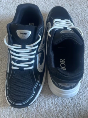 Svarta Dior B30 sneakers - Säljer ett par svarta Dior B30 sneakers med vita och grå detaljer. Skorna har en chunky sula, meshpartier och stora reflekterande Dior-loggor på sidorna. Klassisk snörning med vita skosnören och snygga kontraster. Perfekta för dig som gillar exklusiva och trendiga sneakers.