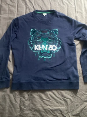 Blå sweatshirt med tiger från Kenzo - Mörkblå sweatshirt från Kenzo med ikoniskt broderat tigerhuvud i turkos, grönt och vitt på bröstet. Stora vita bokstäver med Kenzo Paris under motivet. Rund halsringning och ribbade muddar vid ärmar och nederkant. Perfekt statement-plagg för dig som gillar streetwear. Toppskick förutom att tvättlappen är avklippt pga irritation.