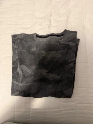Grå camo t-shirt i bomull - Snygg grå t-shirt med camouflage-mönster. Perfekt för en avslappnad streetstyle-look. T-shirten har rund hals och är kortärmad, tillverkad i mjuk bomull för skön komfort.