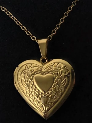 Guldigt hjärthalsband med medaljong Rostfritt Stål  - Snyggt halsband i guldfärgad metall med ett hjärtformat medaljonghänge. Medaljongen har fina ingraverade blommönster och ett slätt hjärta i mitten. Kedjan är tunn och klassisk, och medaljongen går att öppna för att lägga in en liten bild eller minne. Detaljer:  Material: Rostfritt stål. Färg: Guldtonad. Design: Öppningsbar medaljong med graverad framsida. Hänge: 2,9 cm och 2,85 cm. Kedjans längd: 45 cm + 5 cm förlängningskedja. Skick: Nytt