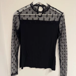 Svart mesh topp med stjärnor - Snygg svart mesh topp med långärmade ärmar och transparent tyg upptill och på ärmarna. Toppen har ett stjärnmönster och en liten knäppning i nacken. Perfekt för dig som vill ha en trendig och unik look.