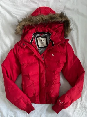 Röd vintage pufferjacka från Abercrombie & Fitch - Snygg vintage röd pufferjacka med huva och fuskpäls från Abercrombie & Fitch. Jackan har ribbade muddar, dragkedja och knappar framtill samt två fickor. Klassisk logga på bröstet och ärmen. Perfekt för kalla vinterdagar. Vintage storlek L passar mer som s-m. Måtten finns. 