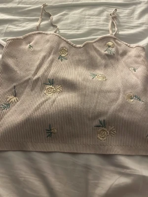 Ljusbeige ribbad  linnetopp - Supersöt ljusbeige linnetopp med tunna axelband och broderade ananasser i gult och grönt. Toppen är ribbad och har en croppad passform som passar perfekt till höga jeans eller kjol. Perfekt för varma dagar och en chill stil.