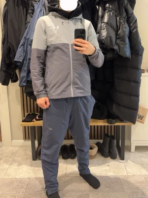 Grå vindjacka från Under Armour - Snygg tvåtonad grå vindjacka från Under Armour med dragkedja framtill och huva. Jackan har en sportig look med logga på bröstet och är tillverkad i lätt, vindtätt material. Perfekt för dig som vill ha en stilren och funktionell jacka till vardags.