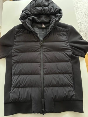 Svart pufferjacka med huva från Duno - Snygg svart pufferjacka från Duno med korta ärmar och vadderad framsida. Jackan har huva, dragkedja framtill och två sidofickor. Perfekt för dig som vill ha en modern och sportig look. Materialet är en mix av polyester och syntet. Original pris är 3899*