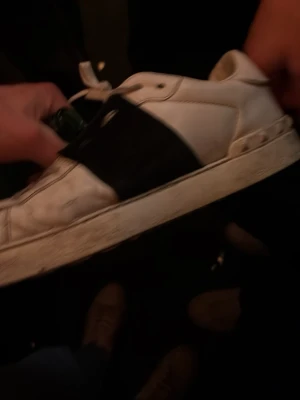 Vita Valentino sneaker från push - Säljer ett par vita sneakers från Valentino med en bred svart rem över mitten och nitar längs hälen. Skorna har rund tå, snörning och platt sula. Perfekta för dig som gillar en clean men edgy stil.