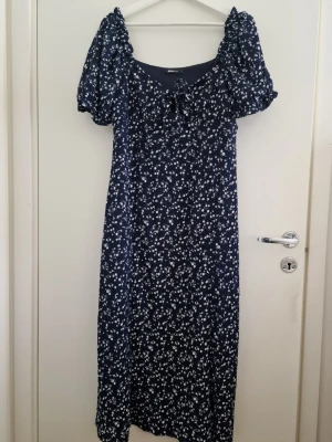 Marinblå blommig midiklänning från Gina Tricot XL - Supersöt marinblå midi sommarklänning från Gina Tricot med vitt blommönster, puffiga kortärmar och v-ringning med knyt fram. Perfekt för sommarens alla tillfällen. Klänningen har en avslappnad passform och är gjord i mjukt, lätt material. Använd en gång, som ny. Storlek XL. Elastisk. 