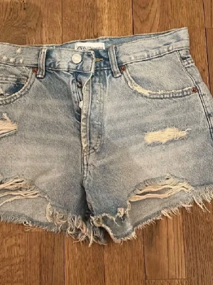  jeansshorts Zara - Säljer ett par ljusblå högmidjade jeansshorts från Zara med slitna detaljer och fransig kant. Klassisk femficksmodell med knappgylf och råa kanter för en avslappnad look.