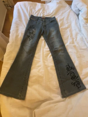 Blå bootcut jeans med broderat mönster - Säljer ett par blå bootcut jeans med svart broderings tryck på båda benen. Dom är midwaist och är precis ner till golvet på mig som är 161 cm. Köpte vintage för 500 kr men passar tyvärr inte mig längre.