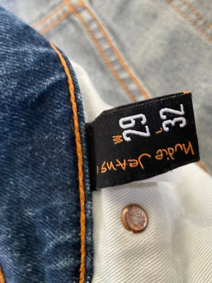 Blå jeans från Nudie Jeans, 29/32 - Säljer ett par klassiska blå jeans från Nudie Jeans i storlek 29/32. Jeansen har snygga kontrastsömmar och ikoniska broderade bakfickor. Perfekta för dig som gillar en tidlös och avslappnad stil.