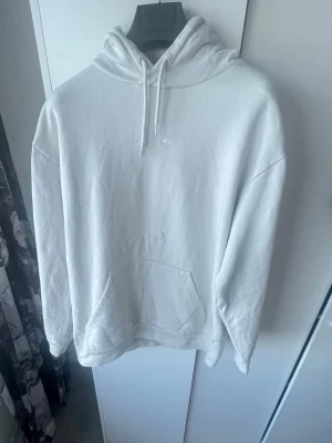 Vit Adidas hoodie  - Säljer en stilren vit hoodie med huva och dragsnören. Hoodien har en klassisk känguruficka framtill och ribbade muddar vid ärmslut och nederkant. Materialet ser ut att vara mjukt och bekvämt, perfekt för en avslappnad look.