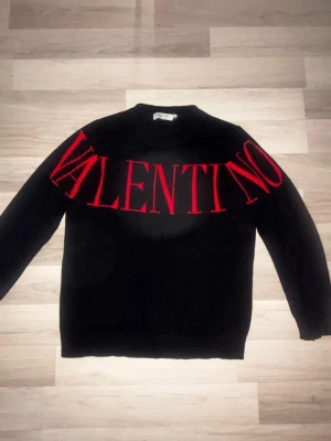 Svart stickad tröja från Valentino - Säljer en svart stickad tröja från Valentino med stor röd logotyp över bröstet och axlarna. Tröjan har rund halsringning och långa ärmar. Perfekt för dig som vill ha en statement piece i garderoben.