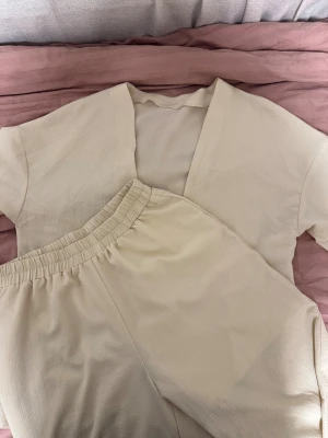 Beige loungset med kimono och byxa - Mysigt loungset i beige med öppen kimono-inspirerad överdel och matchande byxa med resår i midjan. Tillverkat i ett mjukt, strukturerat tyg som känns skönt mot huden. Perfekt för chill hemma eller som sovplagg.