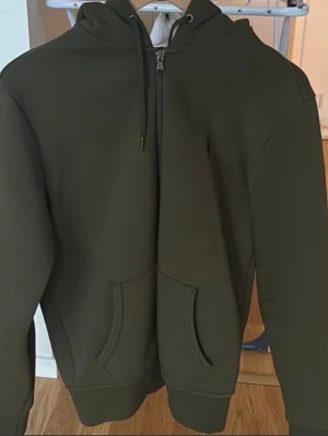 Olivgrön hoodie med dragkedja - Snygg olivgrön hoodie med dragkedja framtill och kängurufickor. Tröjan har en klassisk huva med dragsko och ribbade muddar vid ärmslut och nederkant. Perfekt för en chill och avslappnad stil. Materialet känns mjukt och bekvämt, troligen bomullsblandning.