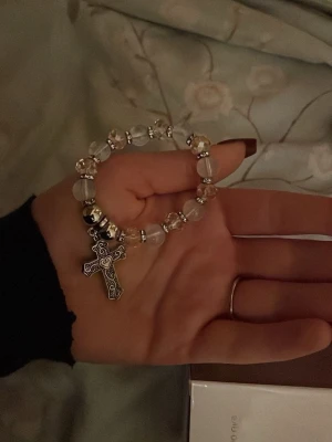 Pärlarmband med korsberlock - Snyggt armband med genomskinliga och silvriga pärlor samt en detaljerad korsberlock i metall. Armbandet har elastisk tråd och pärlorna är runda med metalliska inslag. Perfekt accessoar för dig som gillar unika smycken med personlig touch.