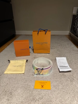 Exklusivt iriserande bälte i rosa/lila – mycket fint skick - Exklusivt och iögonfallande bälte i iriserande rosa/lila ton med elegant silverfärgat spänne. Ett riktigt statement-accessoar som lyfter vilken outfit som helst. Mycket fint skick och varsamt använt. Levereras komplett med originalkartong, dustbag, påse, kvitto och äkthetskort.