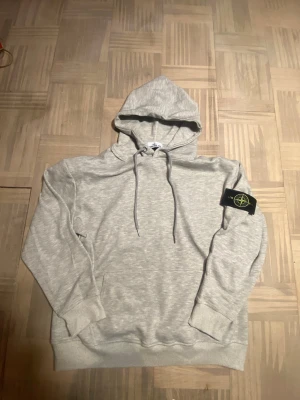 Grå hoodie från Stone Island - Snygg ljusgrå hoodie från Stone Island med klassisk huva och dragsnören. Tröjan har ribbade muddar och den ikoniska Stone Island-loggan på vänster ärm. Tillverkad i mjukt bomullsmaterial som känns skönt mot huden. Perfekt för en avslappnad och trendig look.
