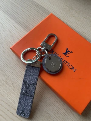  Vuitton nyckelring i monogram - Snygg nyckelring från  Vuitton med klassiskt grått monogrammönster och silverfärgade metalldetaljer. Har både rund ring och karbinhake samt en rund bricka med  Vuitton-logga. Perfekt accessoar till väskan eller nycklarna.