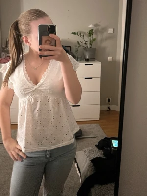 Vit blus med spets och broderi - Söt vit blus med v-ringning, spetskant och broderade hålmönster. Blusen har korta volangärmar och en luftig passform. Perfekt för en fräsch och somrig look.