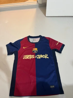 Barça x Cactus Jack matchtröja Nike - Snygg FC Barcelona matchtröja i samarbete med Cactus Jack. Tröjan är röd och marinblå med klubbmärke, Nike-logga och gul text framtill. Tillverkad i lätt och ventilerande material, perfekt för fotboll eller träning.