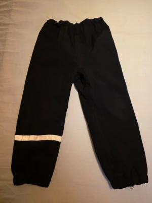 Svarta överdragsbyxor från H&M med reflex - Svarta överdragsbyxor från H&M med resår i midjan. Resoren nedrill har gått sönder. Byxorna har en reflexdetalj runt ena benet för extra synlighet. Perfekta för kalla dagar och vinteraktiviteter. Storlek 104