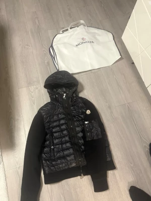 Svart pufferjacka från Moncler - Snygg svart pufferjacka från Moncler med stickade ärmar och huva. Jackan har dragkedja framtill, fickor med knappar och Moncler-logga på bröstet. Perfekt för dig som vill ha en stilren och exklusiv look.