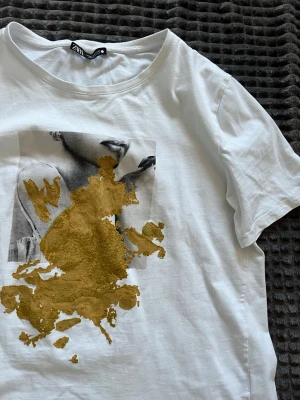 Vit Zara t-shirt med guldtryck  - Vit t-shirt från Zara med ett svartvitt fototryck och en stor abstrakt guldfärgad detalj framtill. Klassisk rund halsringning och korta ärmar. Perfekt för dig som gillar unika prints och vill sticka ut lite extra.Storlek M
